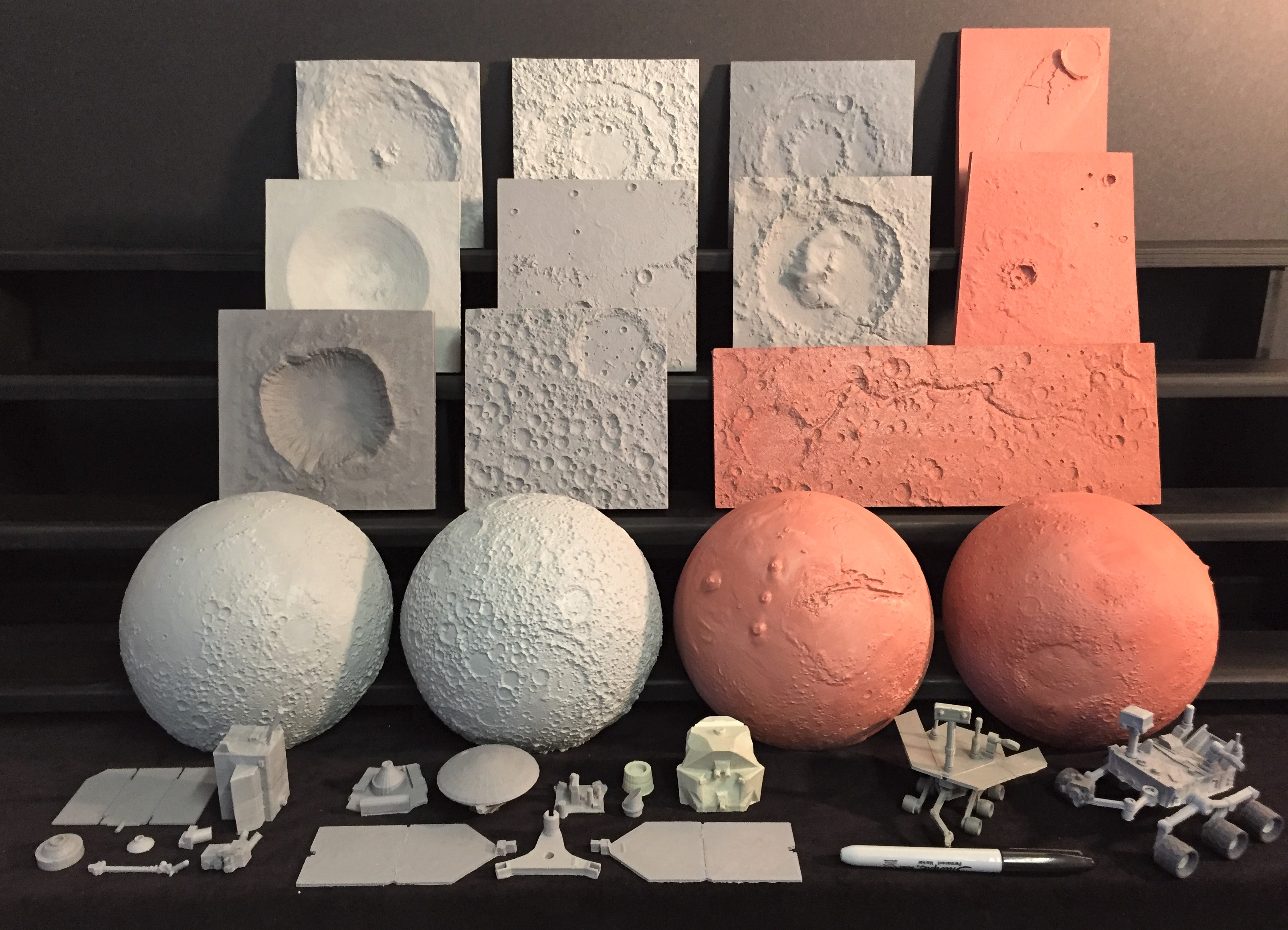 Monolithic 3D models ranging from an impact crater on Earth, five areas of terrain from the Moon, and five areas from Mars.  Hemispherical 3D models of the Moon and Mars are below the monoliths.  Foreground shows 4 different tactile spacecraft kits with Moon and Mars orbiter kits shown as individual pieces and two Mars rovers shown fully assembled.  In the image the two Mars rovers appear to be analyzing a sharpie marker in front of them.