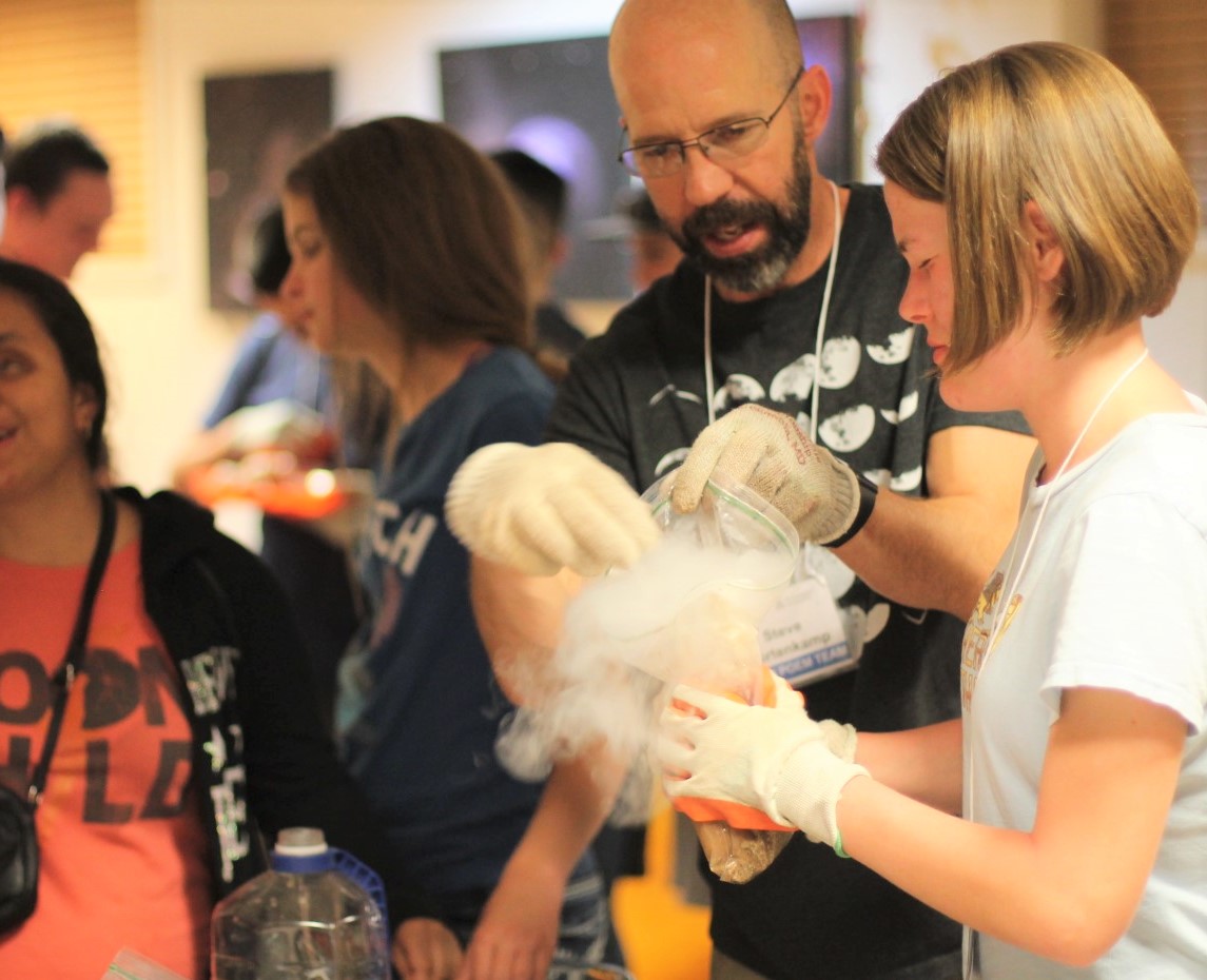 Student and Steve Kortenkamp in the foreground wearing gloves while working with dry ice in comet making activity, with vapor cloud partially obscuring their hands. Image includes additional students working the background. 