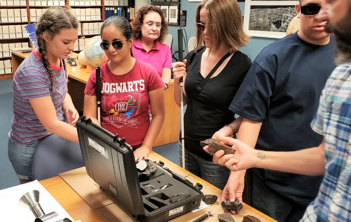Foreground is a table with an open briefcase holding many different meteorites.  Three students are standing at the table and being handed the meteorites.  Irene Topor is in the background looking on.