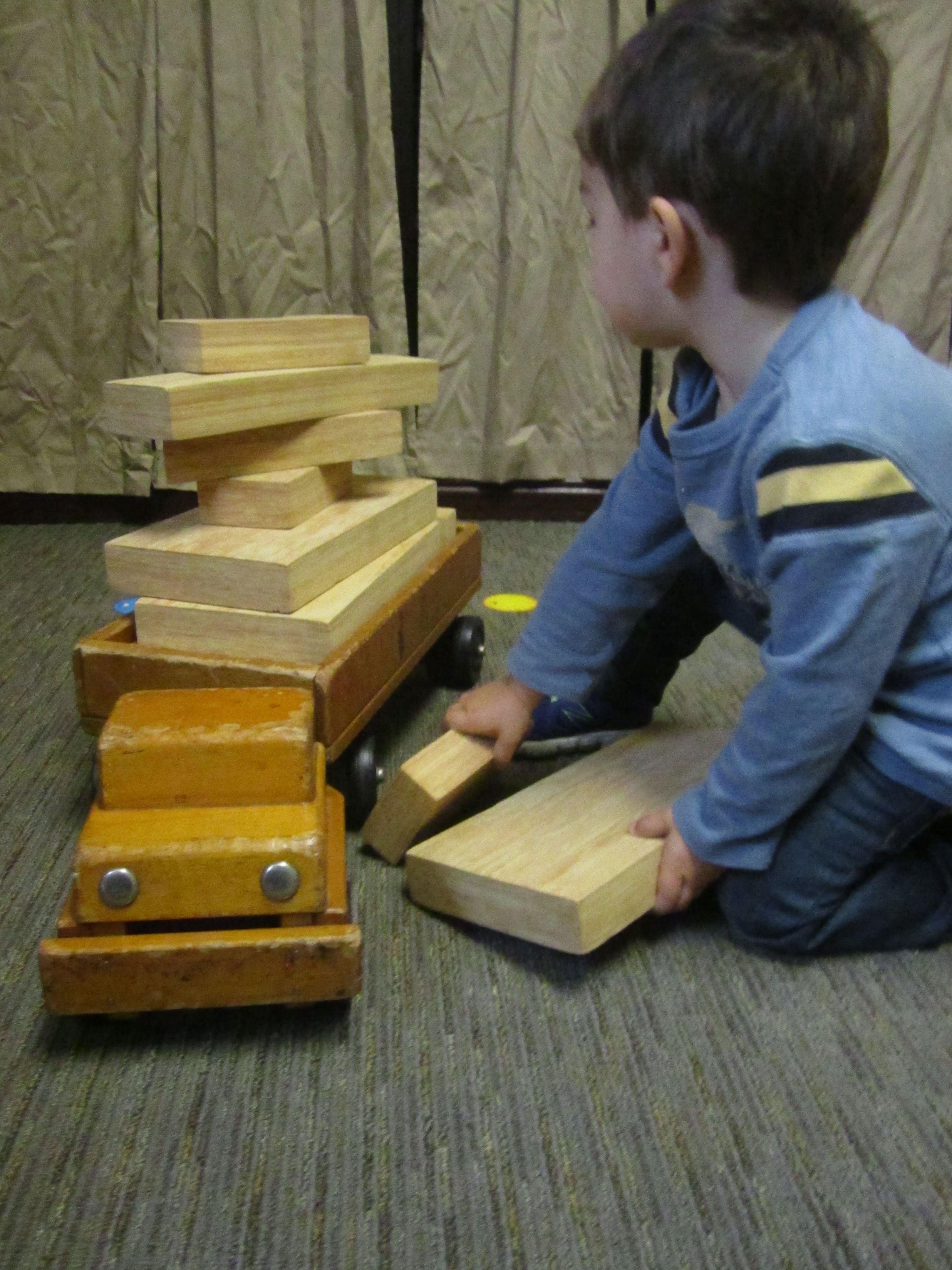 Child stacking wooden unit blocks on top of a wooden truck for transport in a classroom.