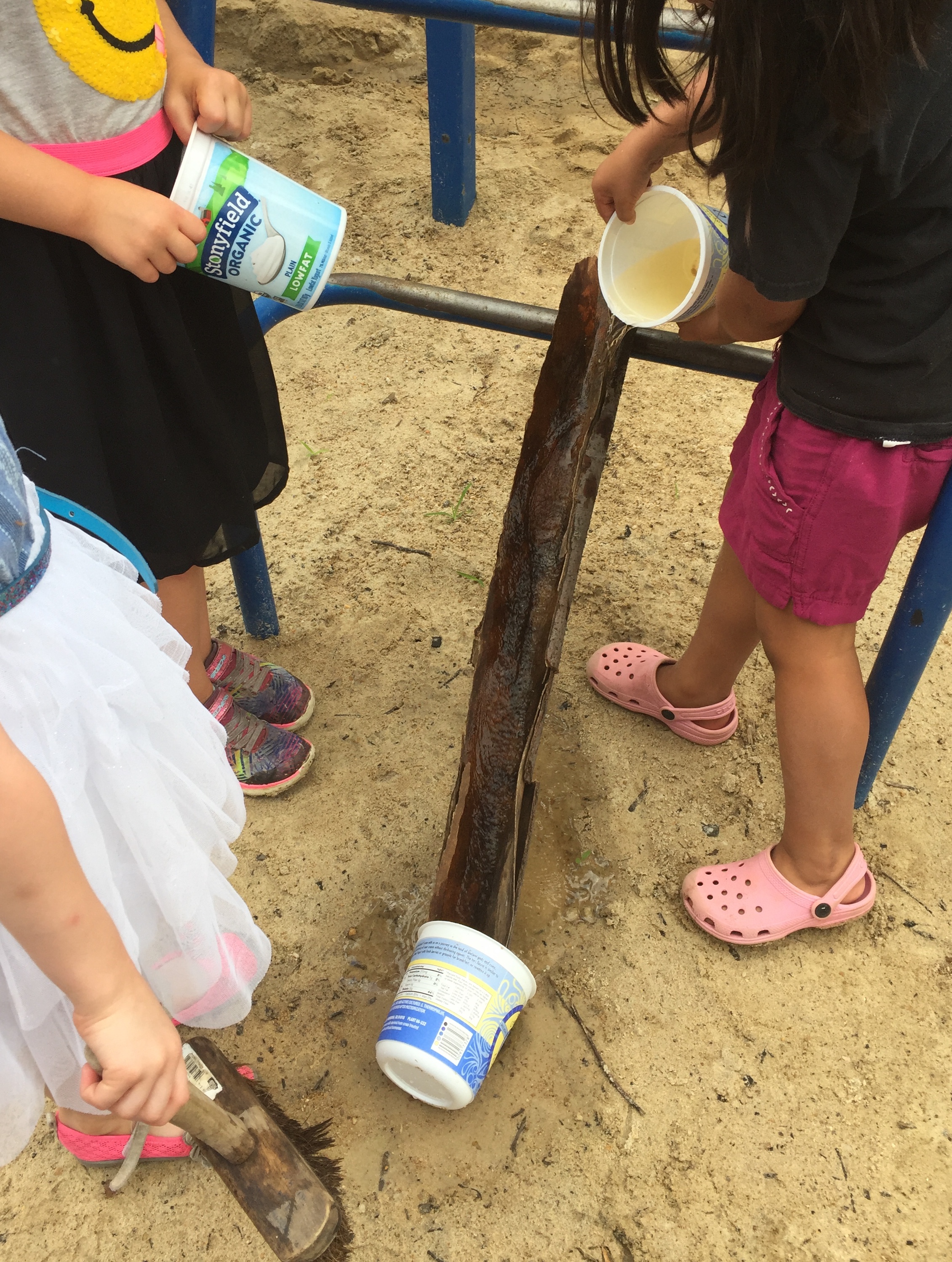 Other children join in the activity, pouring water onto the trough-like piece of tree bark.