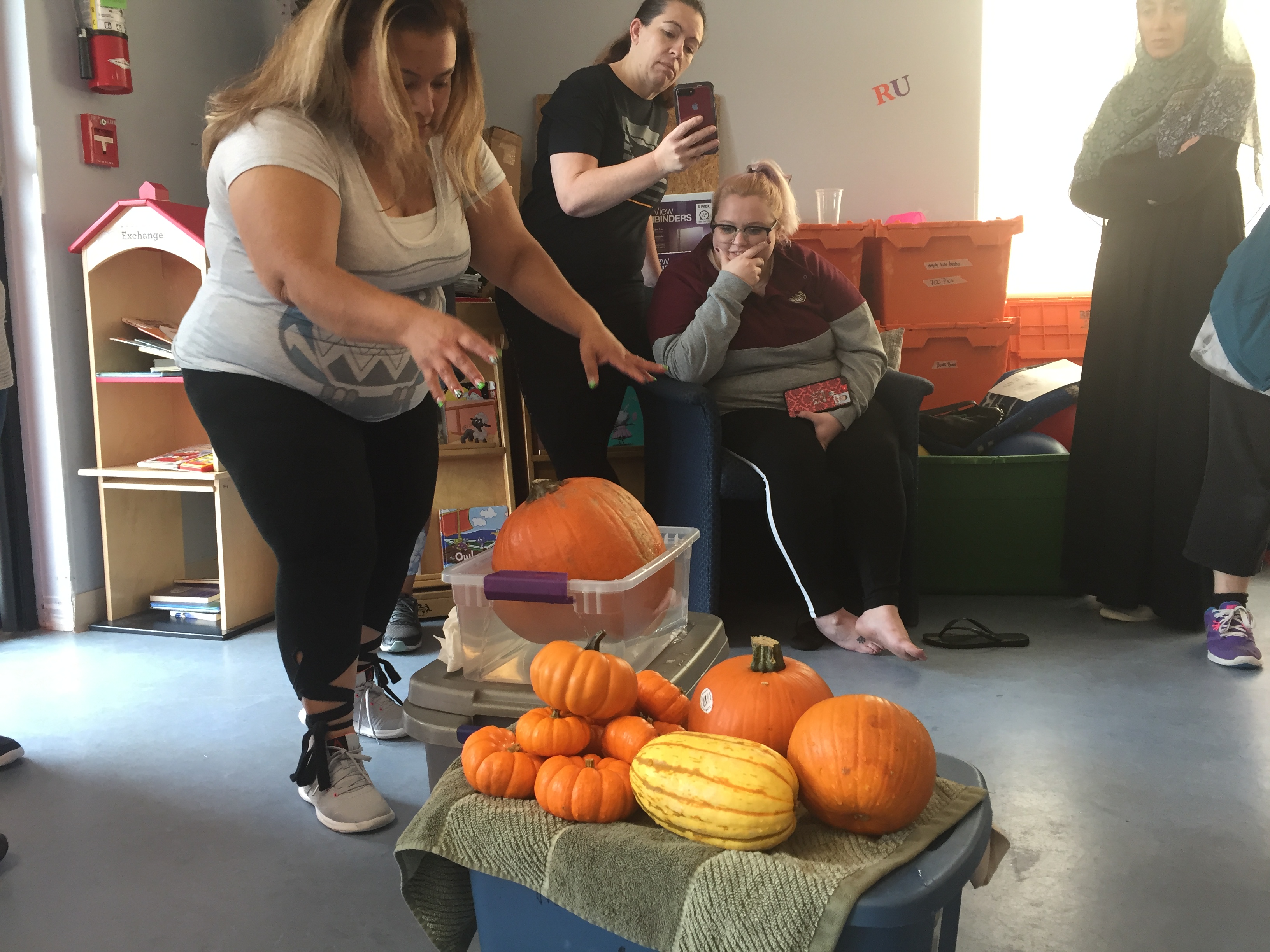 Teacher putting a large pumpkin into a tub of water. Next to it are many smaller pumpkins.