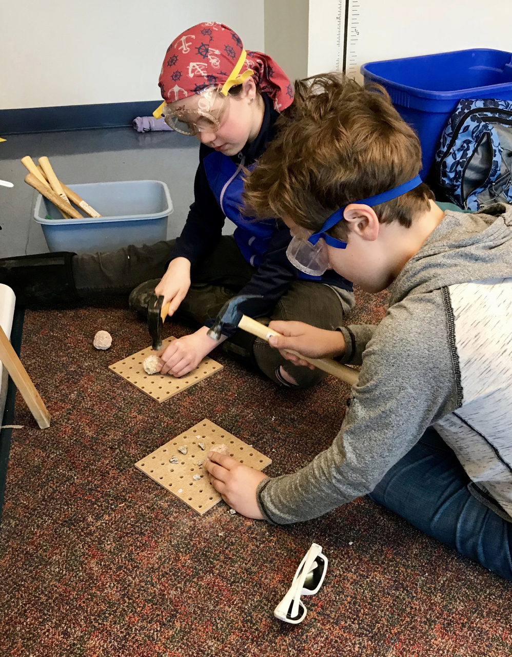 Elementary students wearing goggles using hammers to break open rock.