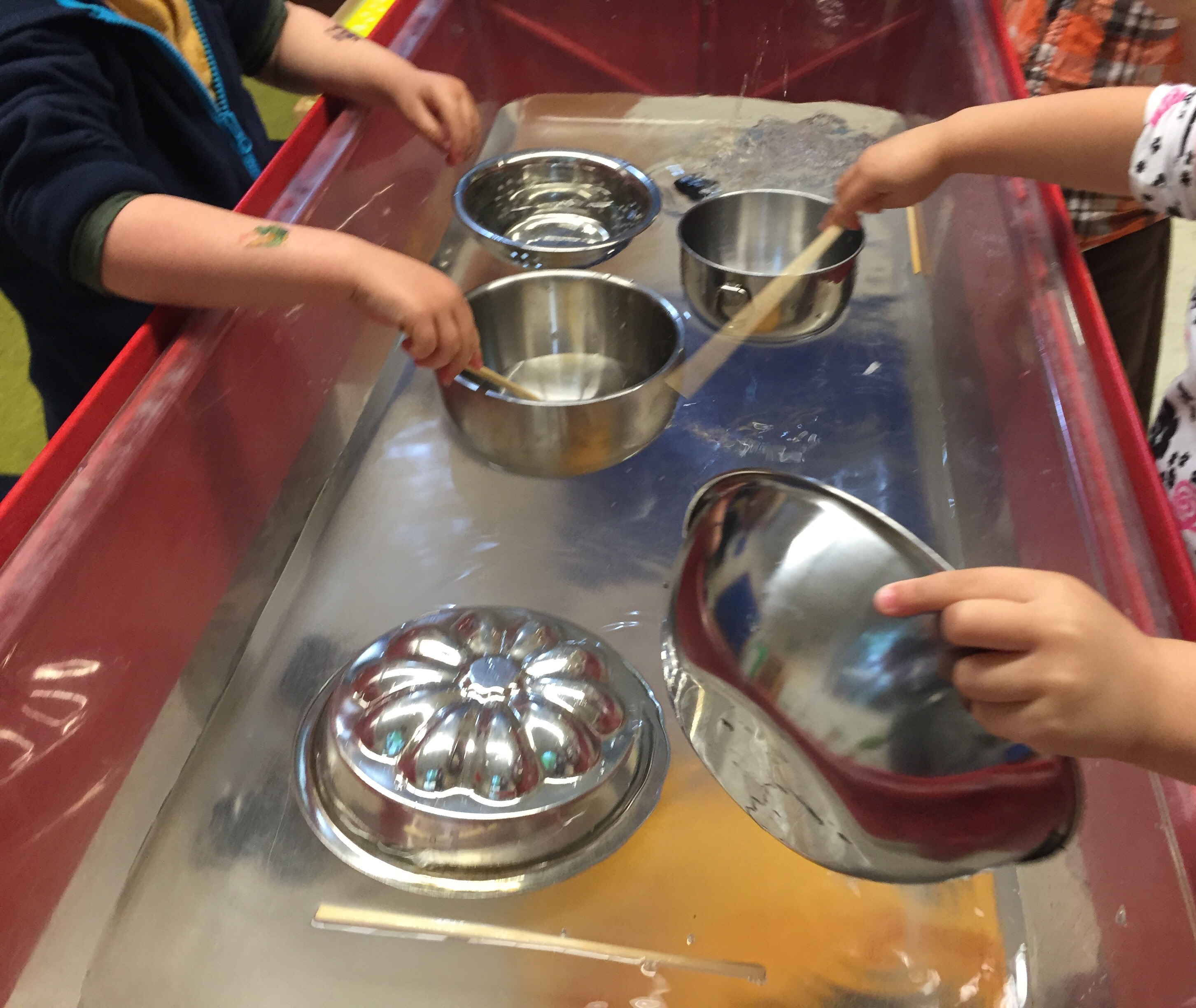 Children play in a water table with water and metal bowls, experiencing sound and wetness.