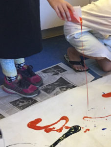 Child pouring paint from waist height on to a sheet on the floor.
