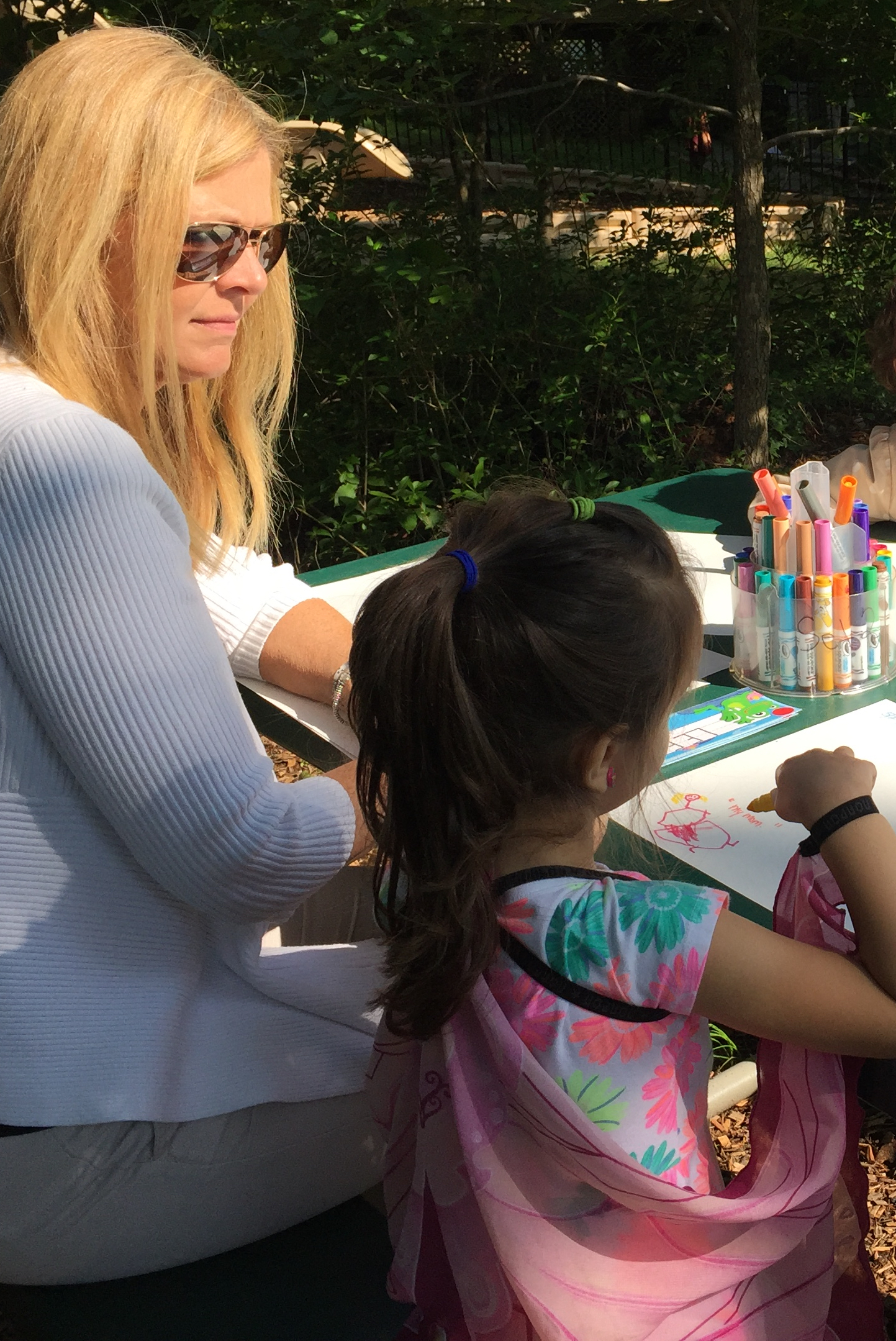 Teacher taking anecdotal notes as child sitting next to her at a picnic table talks about her drawing.