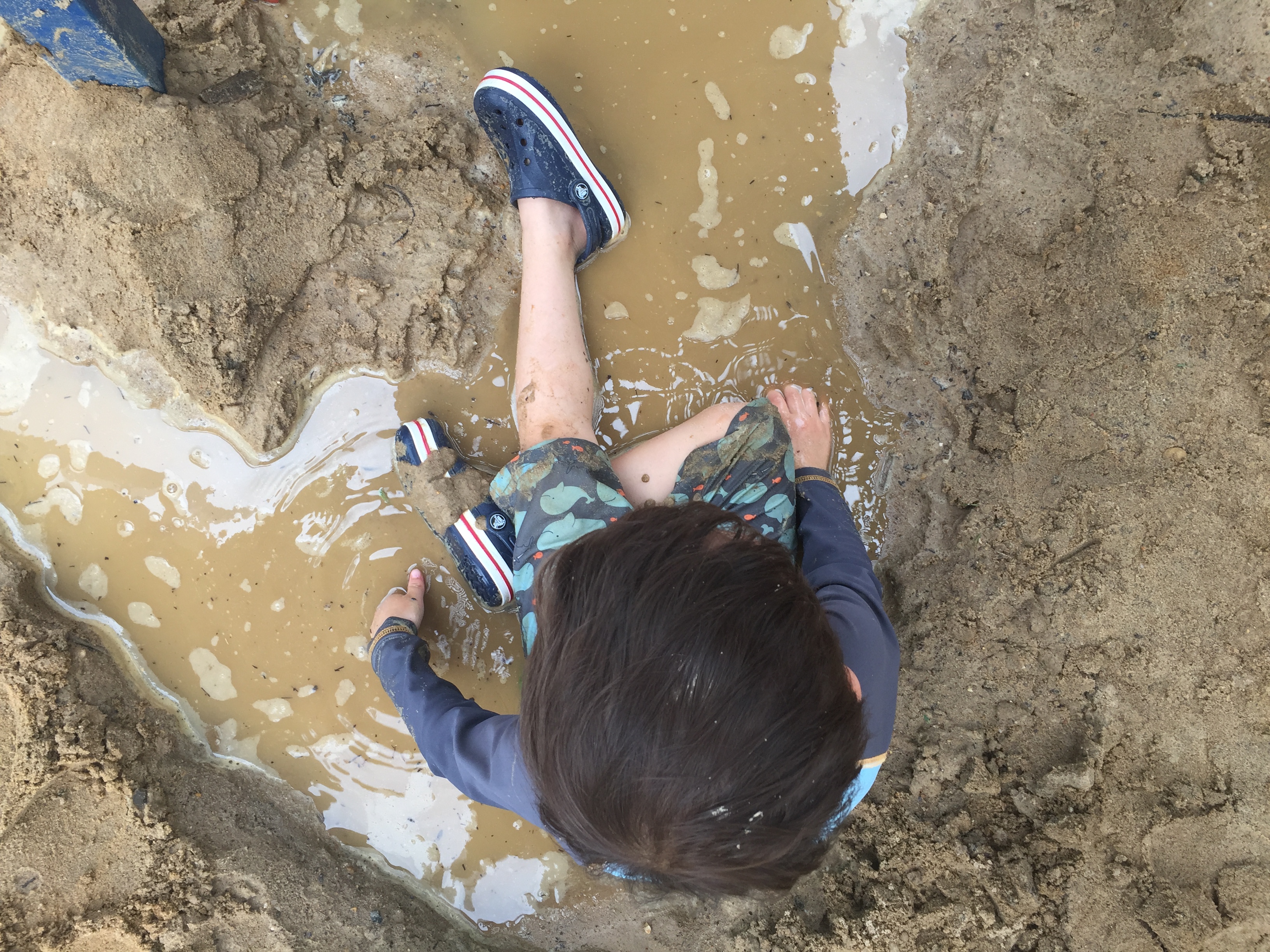 Child in water shoes and suit sitting in a puddle of water in the sand.