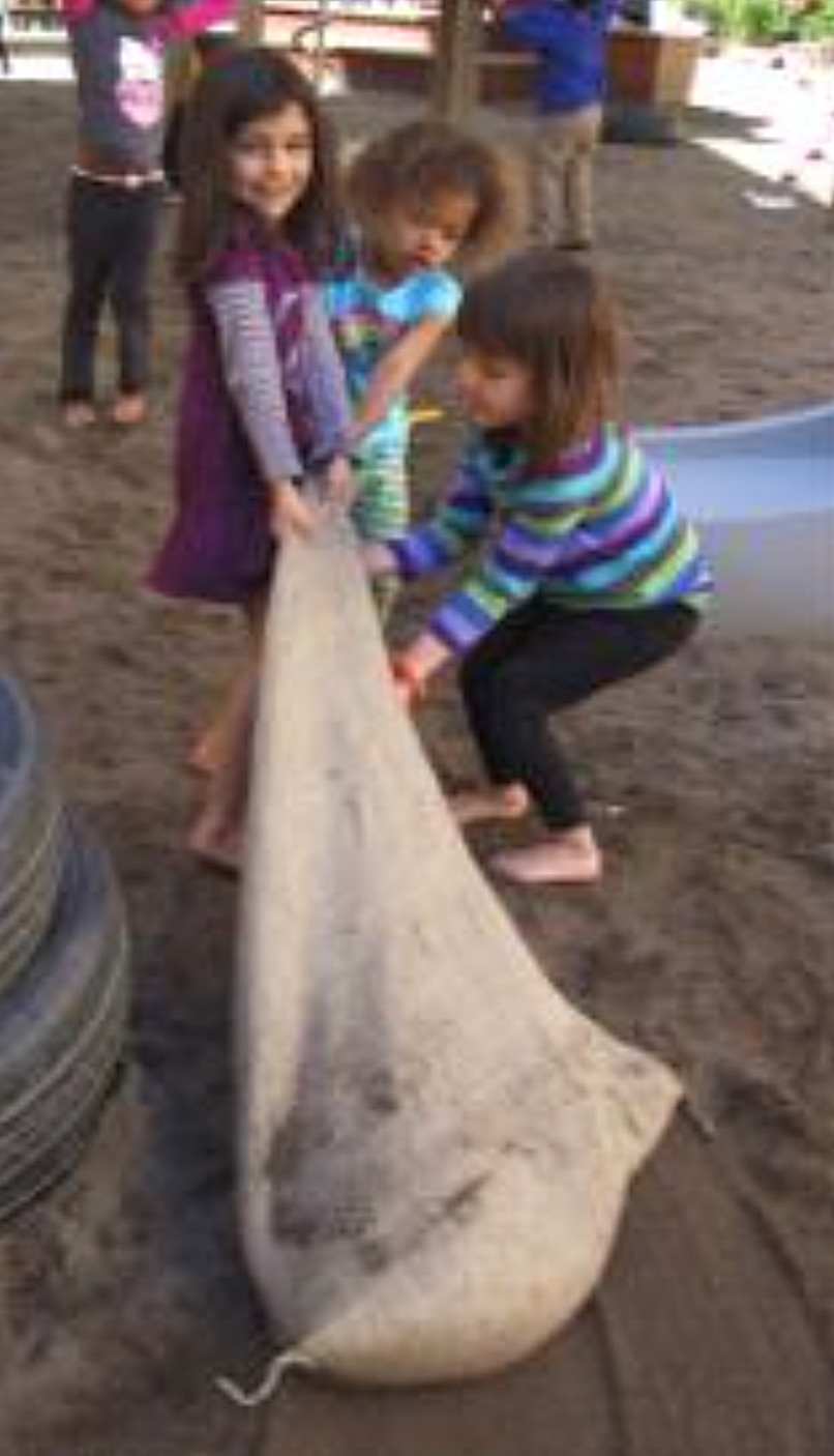 3 preschool girls pulling a heavy sack across a sandy playground