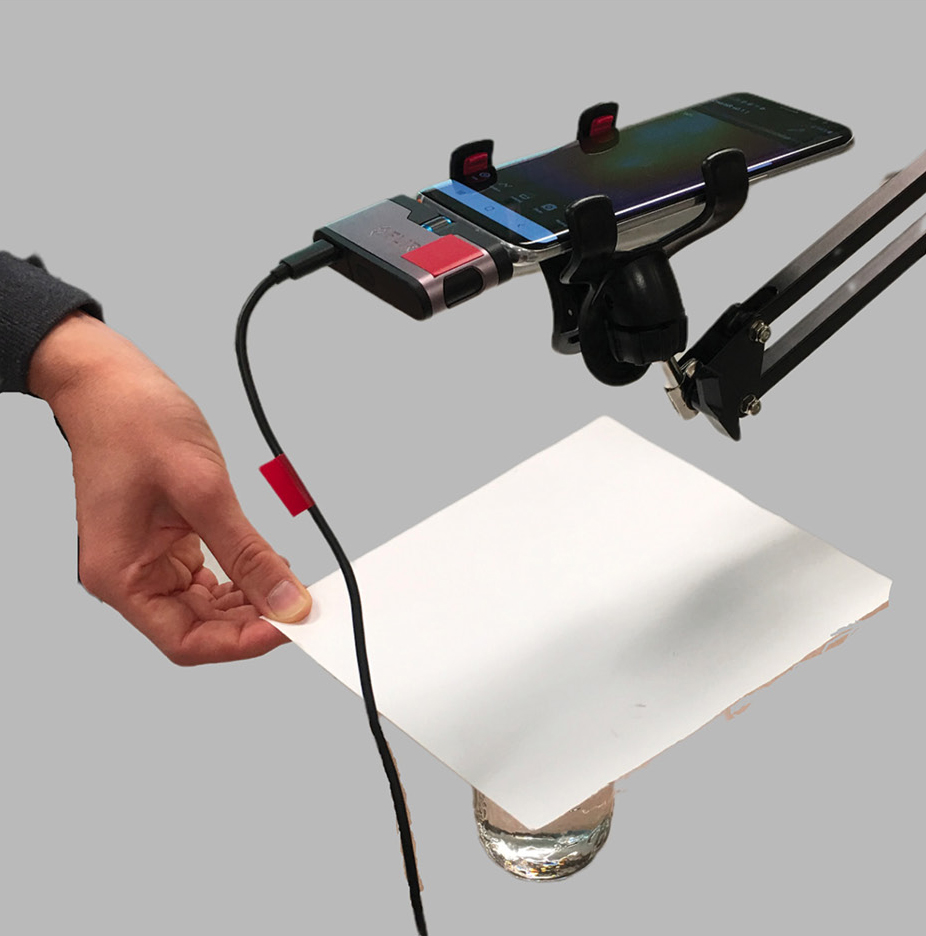 A typical Telelab setup consisting of a smartphone, a sensor module attached to the phone, a phone stand, and the experiment in question. This image shows a demonstration of heating due to condensation of H2O molecules using a piece of paper over a cup of water (Xie 2011).